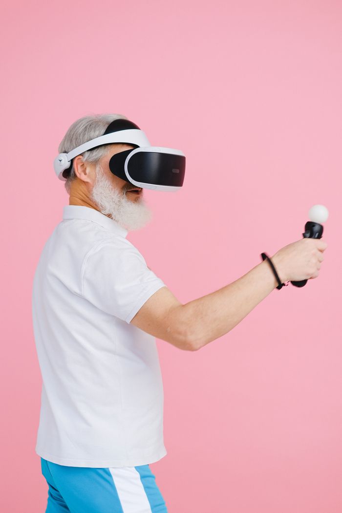 Elderly man using VR headset and controller on pink background.