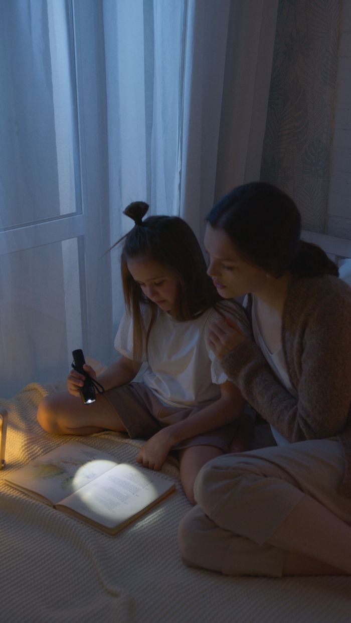 A mother and daughter enjoy a nighttime reading session together by flashlight in a cozy indoor setting.
